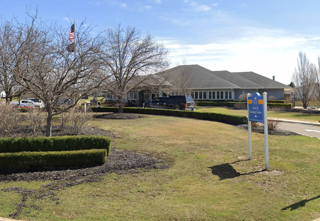 A building with a sales and leasing center sign, surrounded by trimmed hedges and bare trees, under a clear blue sky with an American flag visible.