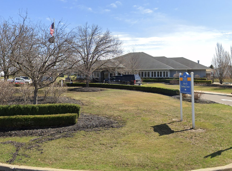 A building with a sales and leasing center sign, surrounded by trimmed hedges and bare trees, under a clear blue sky with an American flag visible.