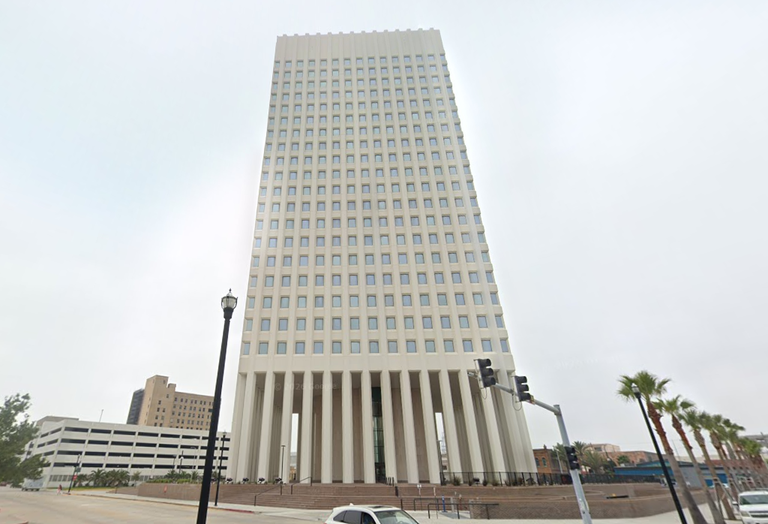 Tall, rectangular white building with uniform grid windows, standing amidst a street with palm trees and traffic lights on a cloudy day.