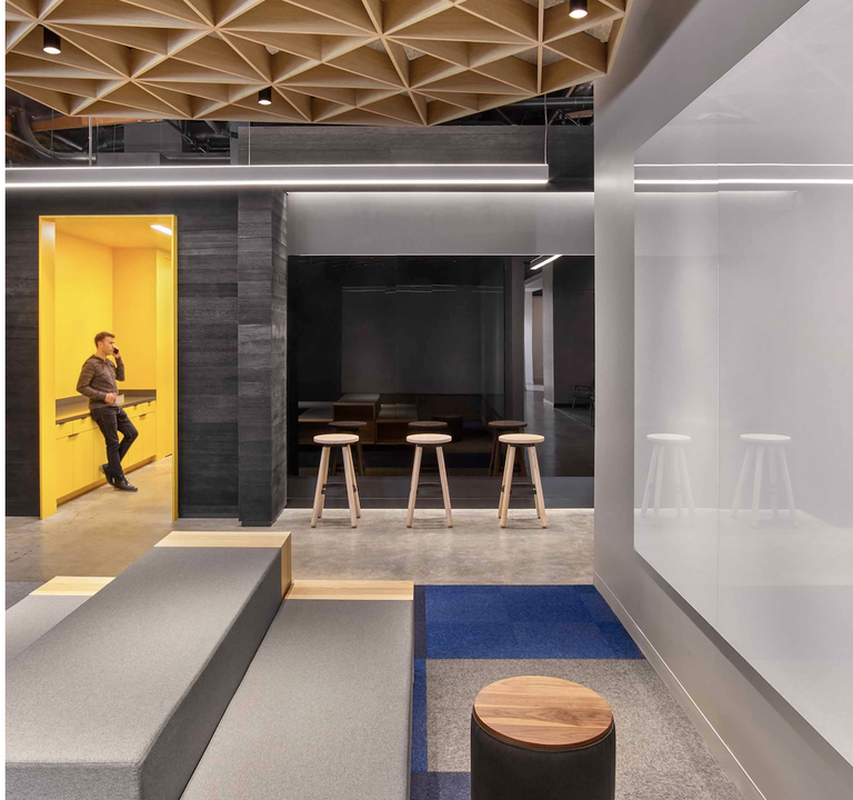 Modern office space with a man in a yellow alcove, wooden stools, geometric ceiling, and grey seating, creating a vibrant and contemporary atmosphere.