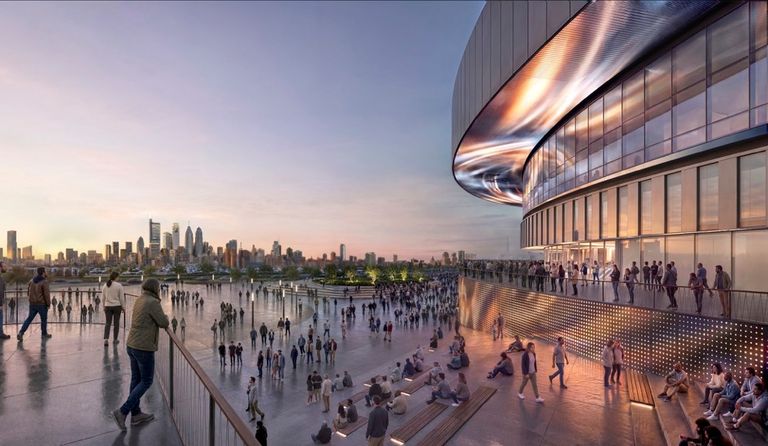 People gather on a large terrace overlooking a city skyline at sunset, near a modern, illuminated building.