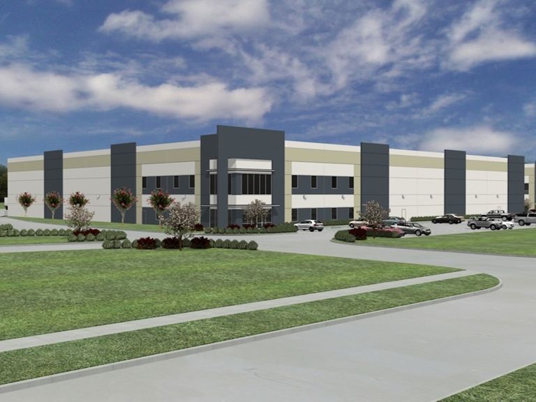 A modern industrial building with a large parking area, surrounded by landscaped greenery under a partly cloudy blue sky.