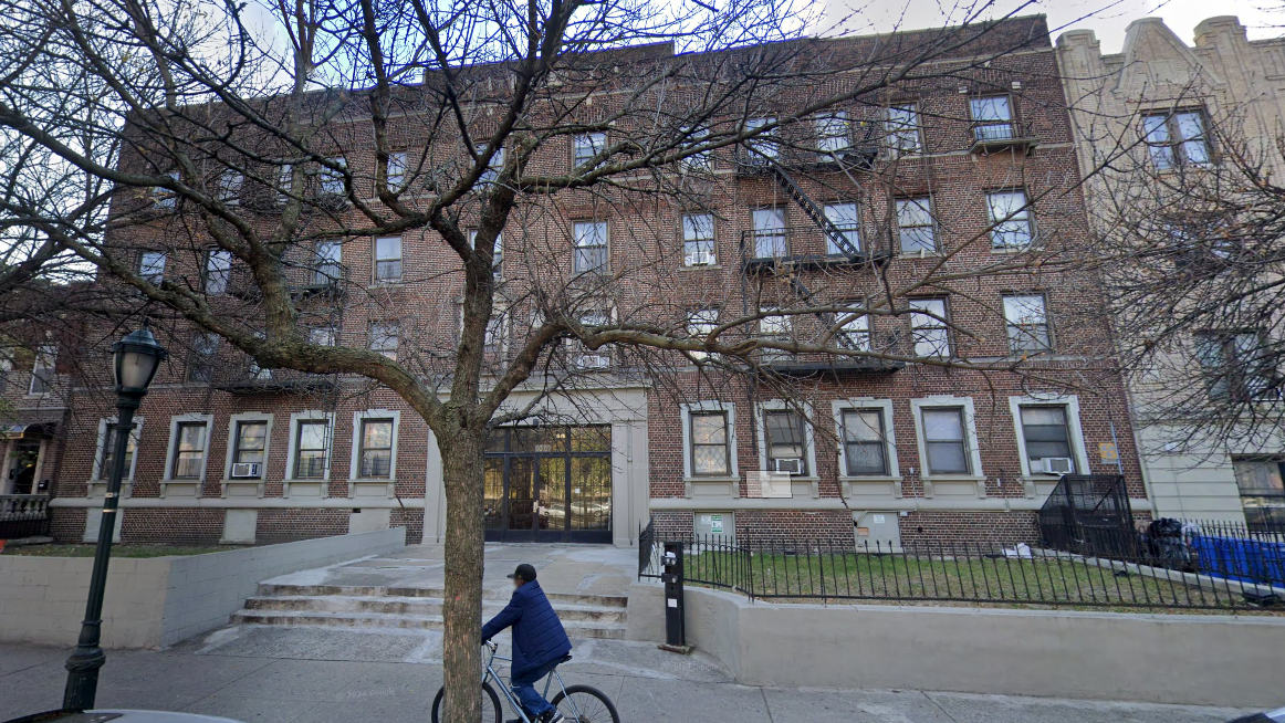 A person rides a bicycle on the sidewalk in front of a large brown brick apartment building with multiple windows and a tree in the foreground.
