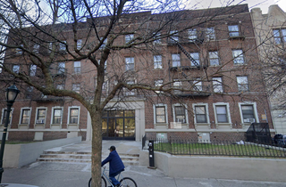 A person rides a bicycle on the sidewalk in front of a large brown brick apartment building with multiple windows and a tree in the foreground.