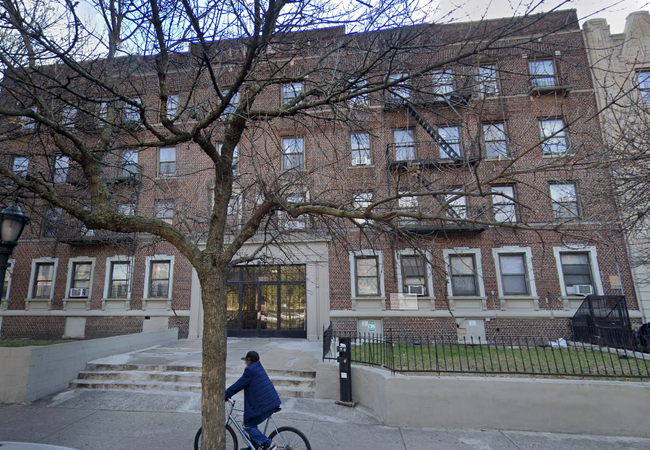 A person rides a bicycle on the sidewalk in front of a large brown brick apartment building with multiple windows and a tree in the foreground.