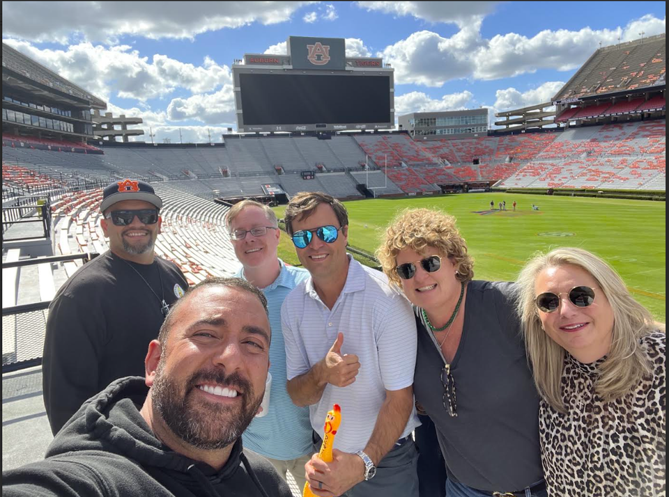 A group of people smiling and posing on a sunny day in a large, empty sports stadium with a clear sky overhead.