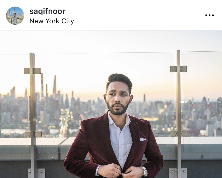 Man in a maroon blazer stands on a rooftop with a glass barrier, city skyline and sunset in the background.