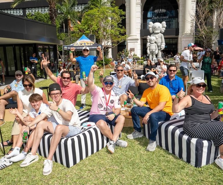 A group of people sitting on striped cushions on grass, smiling and posing with drinks, with a large cartoonish statue in the background.