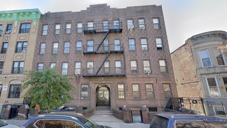 Four-story brick apartment building with fire escapes, surrounded by similar structures. Cars parked in front and a tree near the entrance.