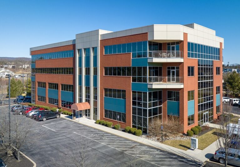 A modern, four-story office building with large windows, red brick and teal paneling, surrounded by a parking lot and landscaped bushes.