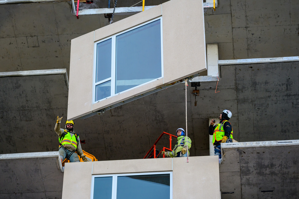 Construction workers guiding a prefabricated wall section with windows into place on a building site.