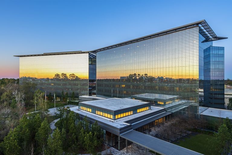 Modern glass building reflecting a sunset, surrounded by lush green trees, under a clear blue sky.