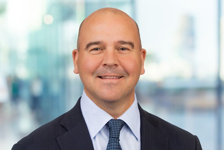 A bald man in a dark suit and tie smiles in a bright, blurred office setting.