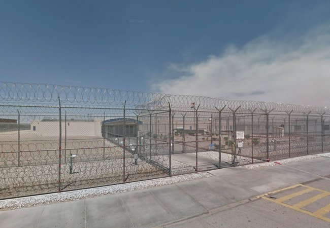 A secure facility with high barbed-wire fencing and a locked gate, set against a clear blue sky and concrete pavement.