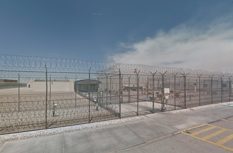 A secure facility with high barbed-wire fencing and a locked gate, set against a clear blue sky and concrete pavement.