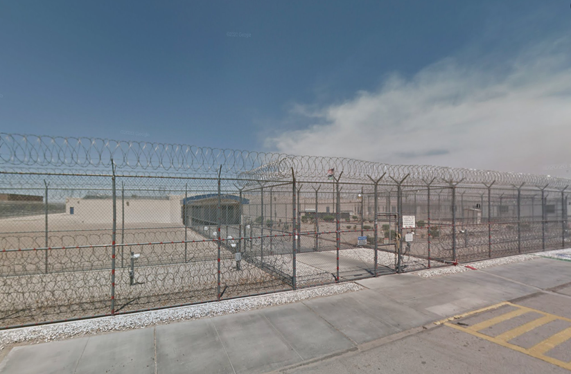 A secure facility with high barbed-wire fencing and a locked gate, set against a clear blue sky and concrete pavement.