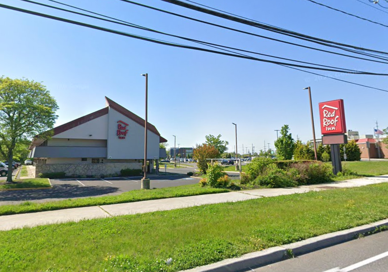 Red Roof Inn building with a large sign, surrounded by greenery, under a clear blue sky. Parking lot and sidewalk visible in foreground.