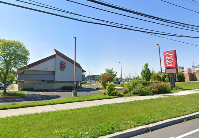 Red Roof Inn building with a large sign, surrounded by greenery, under a clear blue sky. Parking lot and sidewalk visible in foreground.
