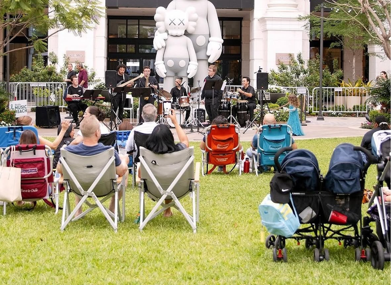 Outdoor concert with people seated on lawn chairs, a band playing on stage, and a large gray sculpture in the background.