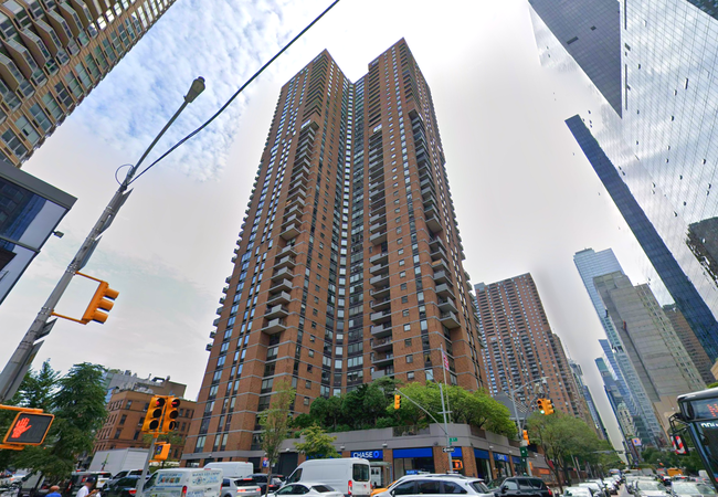 Tall brown brick apartment building with balconies, surrounded by traffic and skyscrapers, under a cloudy sky.