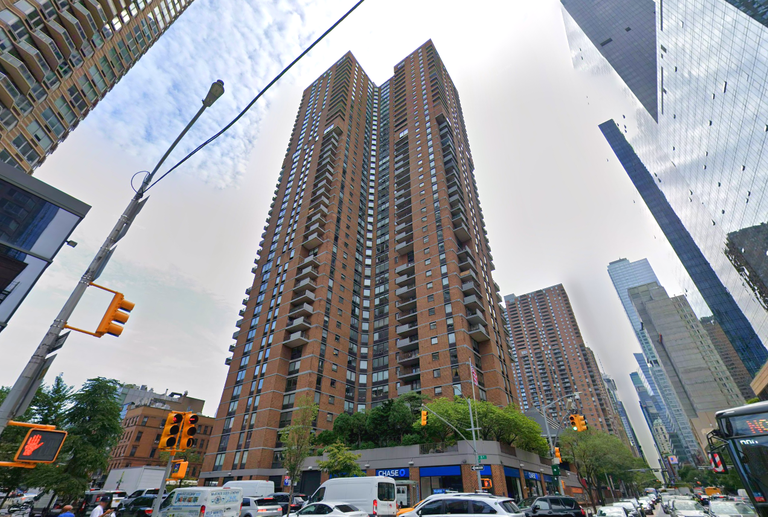 Tall brown brick apartment building with balconies, surrounded by traffic and skyscrapers, under a cloudy sky.