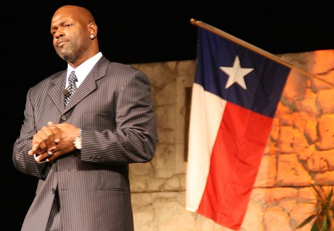 Emmitt Smith stands with clasped hands, next to a Texas flag, against a stone backdrop.