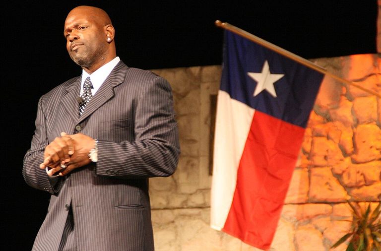 Emmitt Smith stands with clasped hands, next to a Texas flag, against a stone backdrop.