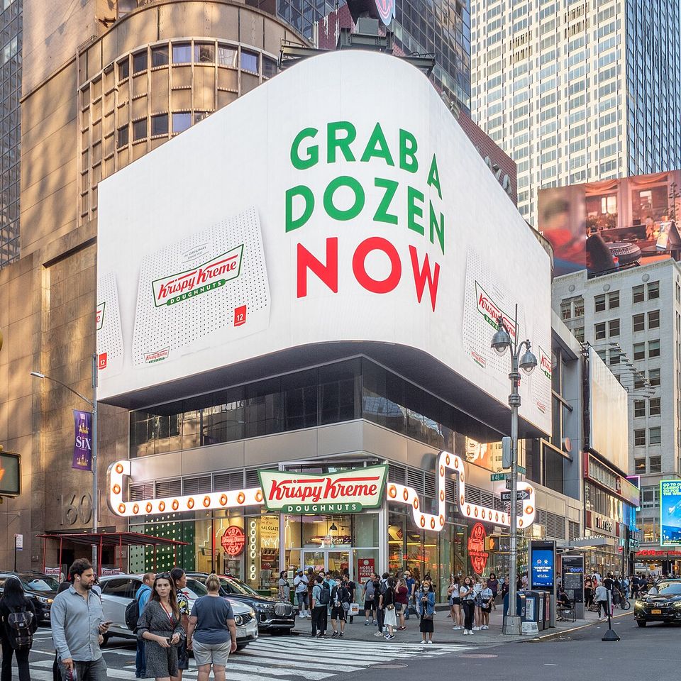 Krispy Kreme store in a busy cityscape with a large billboard saying “Grab a dozen now” and people gathering outside.