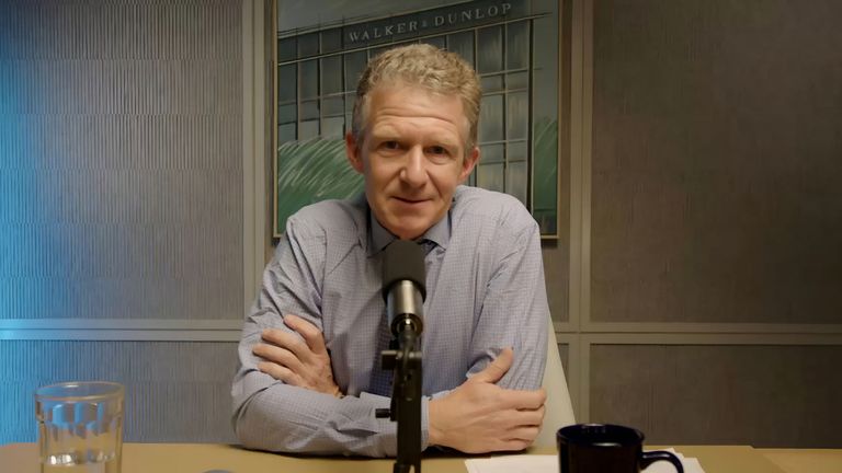 A man in a blue shirt sits at a table with crossed arms, speaking into a microphone with a backdrop of a building photo behind him.