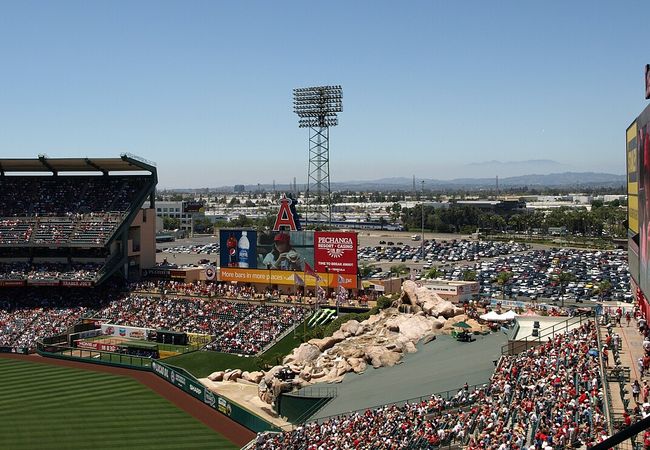 Angels baseball stadium with a packed crowd, large scoreboard, outfield rock feature, and distant view of mountains under a clear blue sky.