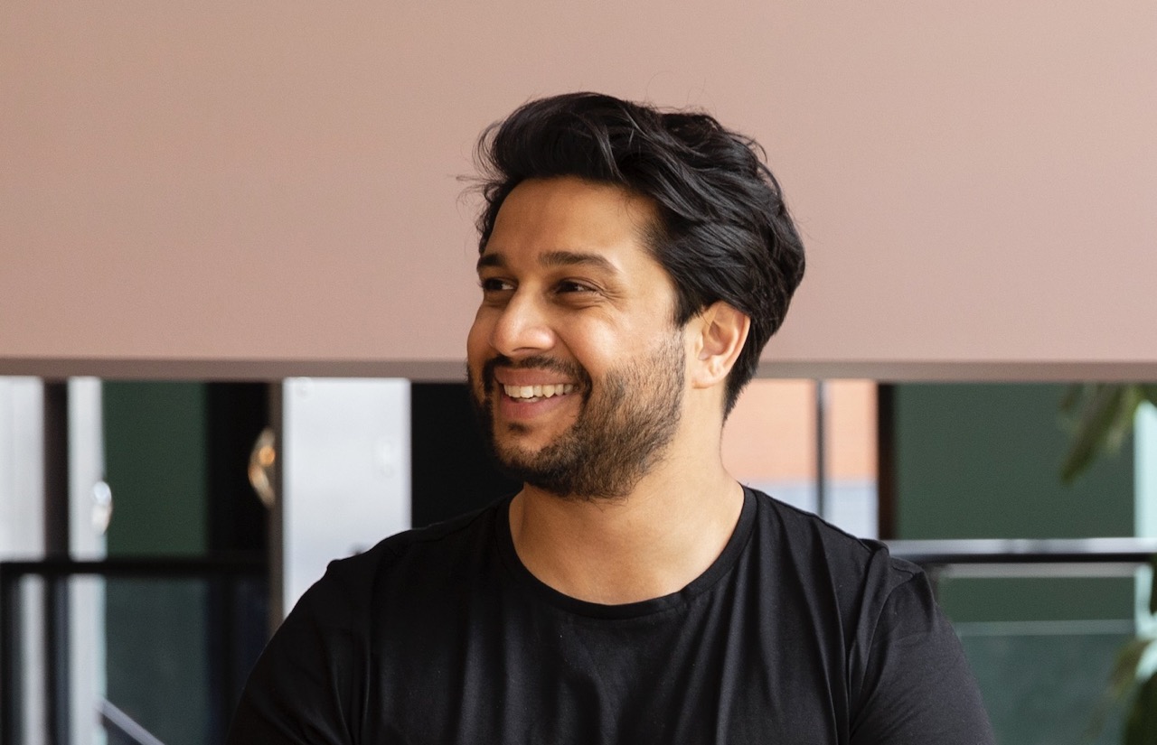 Smiling man with a beard and short black hair wearing a black shirt, seated against a blurred indoor background.