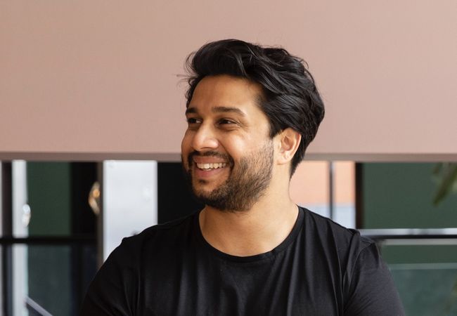 Smiling man with a beard and short black hair wearing a black shirt, seated against a blurred indoor background.