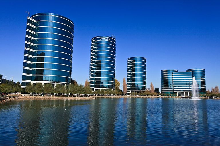Series of modern, cylindrical glass office buildings reflecting in a tranquil lake, with a clear blue sky and fountain in the background.
