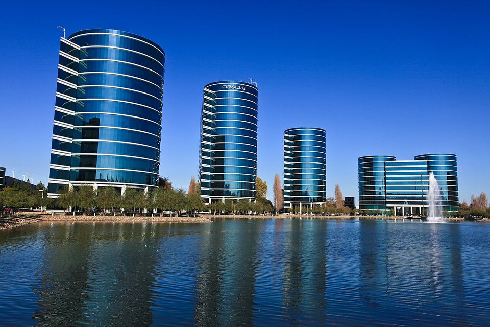 Series of modern, cylindrical glass office buildings reflecting in a tranquil lake, with a clear blue sky and fountain in the background.