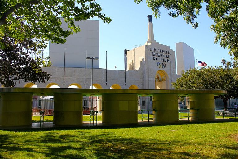 The Los Angeles Memorial Coliseum facade and entrance, surrounded by greenery, with an Olympic symbol visible.