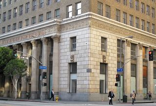 A historic building with ornate columns at the corner of Olive and 7th streets. People walk by, and traffic lights control the intersection.