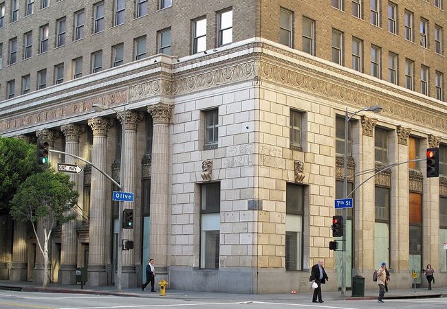 A historic building with ornate columns at the corner of Olive and 7th streets. People walk by, and traffic lights control the intersection.