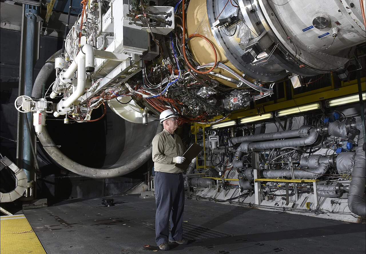 A person wearing a hard hat and gloves examines a large industrial machine or engine in a facility, with numerous wires and pipes visible.