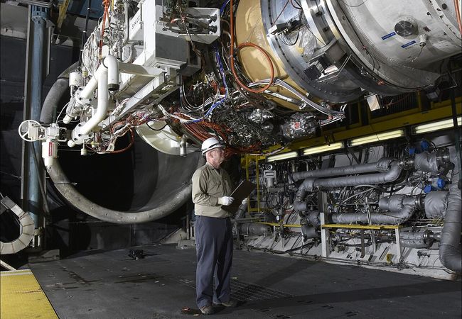 A person wearing a hard hat and gloves examines a large industrial machine or engine in a facility, with numerous wires and pipes visible.