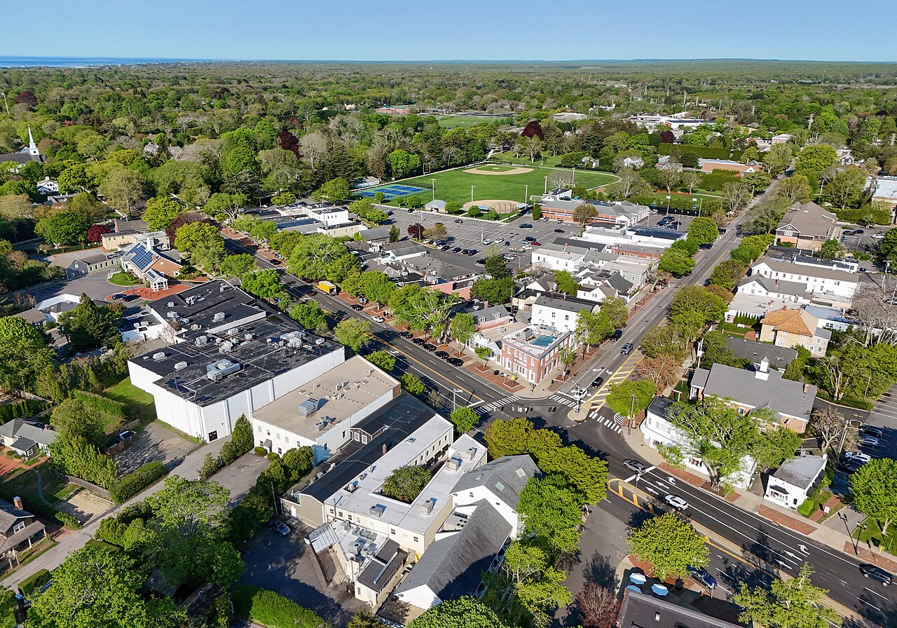 Aerial view of a small town with tree-lined streets, residential and commercial buildings, parking lots, and a sports field, surrounded by lush greenery.