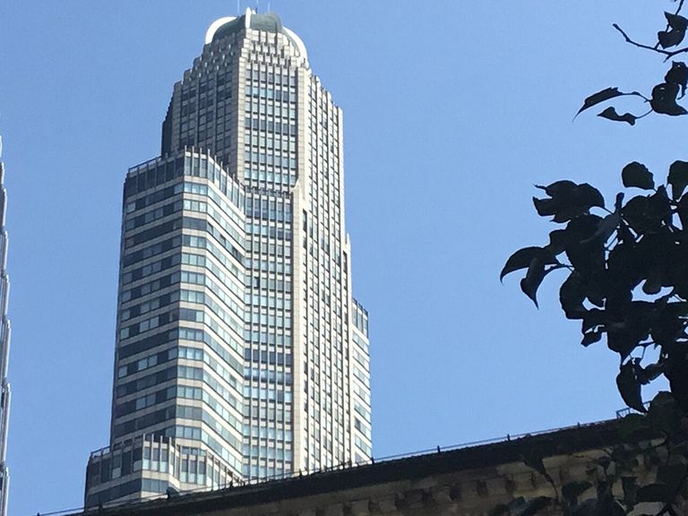 A modern skyscraper with a tiered facade under a clear blue sky, with tree branches partially framing the right side.