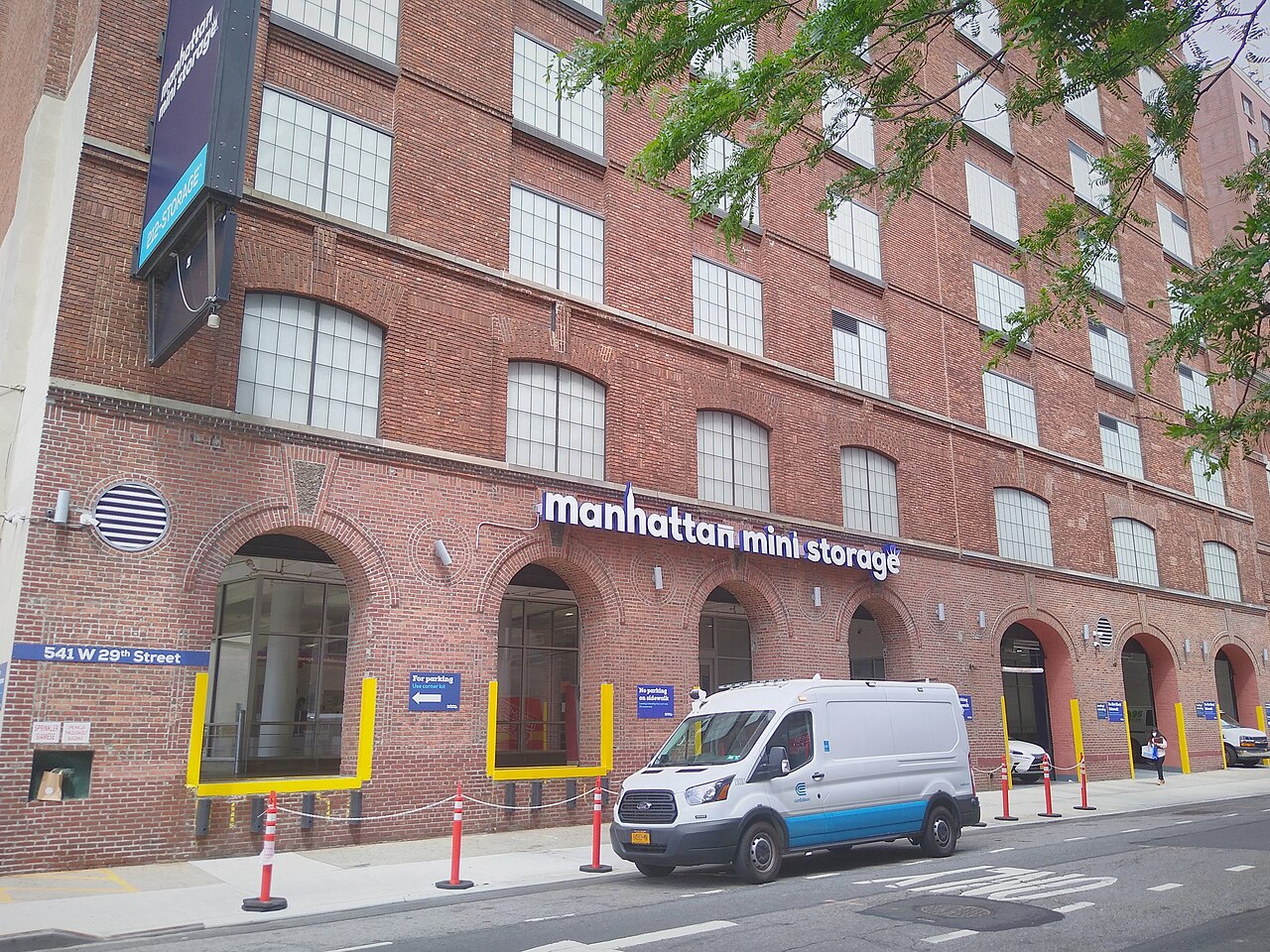 Brick building with large windows labeled "Manhattan Mini Storage." A white van is parked in front, and a person walks on the sidewalk.