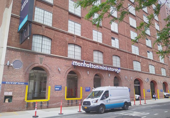 Brick building with large windows labeled "Manhattan Mini Storage." A white van is parked in front, and a person walks on the sidewalk.