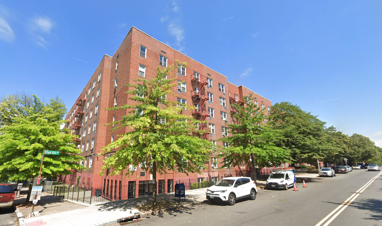 A six-story brick apartment building with fire escapes and trees in front, located on a sunny street with parked cars and a mail drop box.