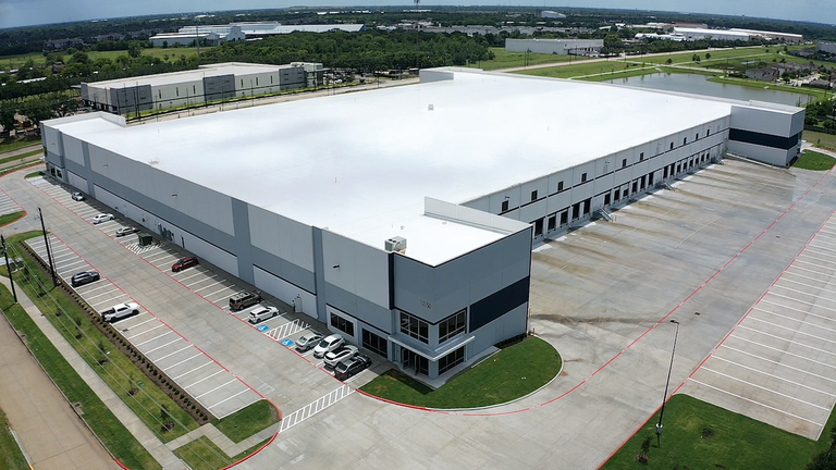 Aerial view of a large industrial warehouse with a white roof, surrounded by parking spaces and green grassy areas, under a clear sky.
