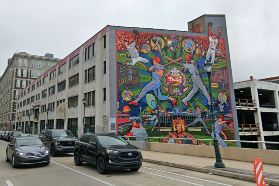 Colorful mural on a building wall depicting baseball players in action, with cars parked on the adjacent street below a cloudy sky.