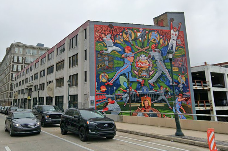 Colorful mural on a building wall depicting baseball players in action, with cars parked on the adjacent street below a cloudy sky.