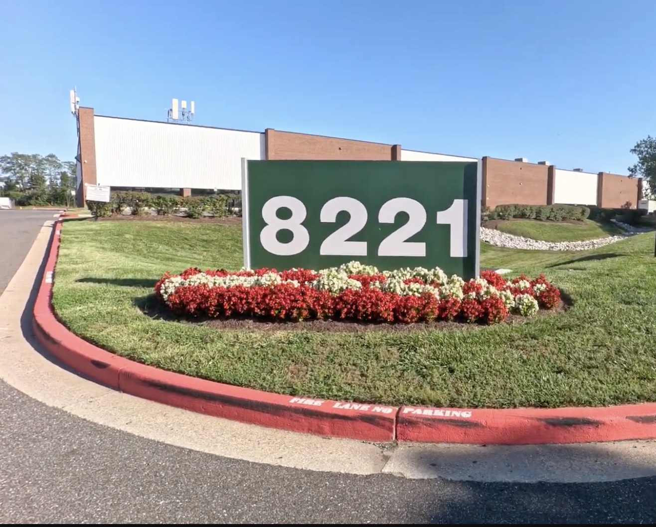 A green sign with the number 8221 stands on a landscaped area with red and white flowers, in front of a large commercial building.