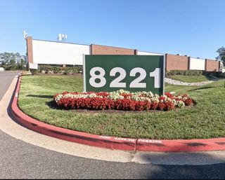 A green sign with the number 8221 stands on a landscaped area with red and white flowers, in front of a large commercial building.