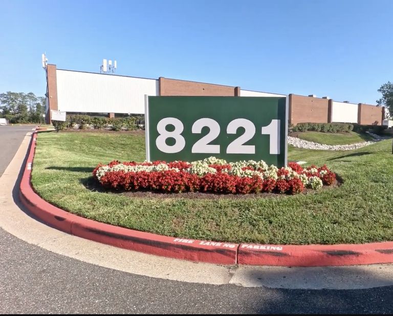 A green sign with the number 8221 stands on a landscaped area with red and white flowers, in front of a large commercial building.